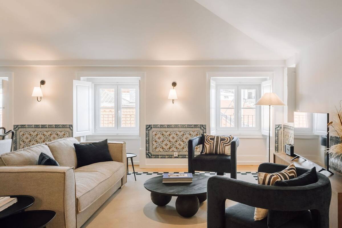 Ein Wohnzimmer mit einer Couch, Sesseln, einem Couchtisch und einer Lampe. Es gibt Fenster mit weißen Rahmen und dekorative Wandverkleidungen.