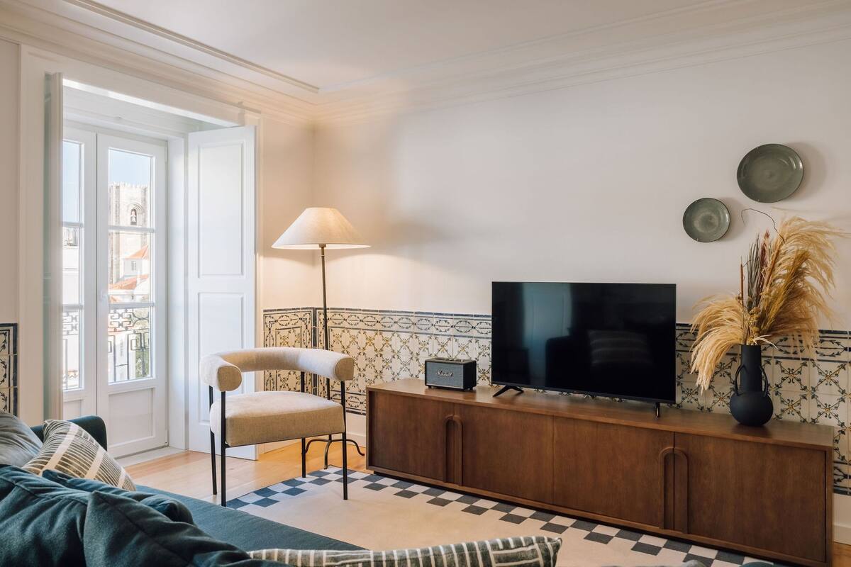 Ein Wohnzimmer mit einer Couch, einem Sessel, einem Fernseher auf einer Holzkommode, einer Lampe und einer Vase mit Trockenpflanzen.