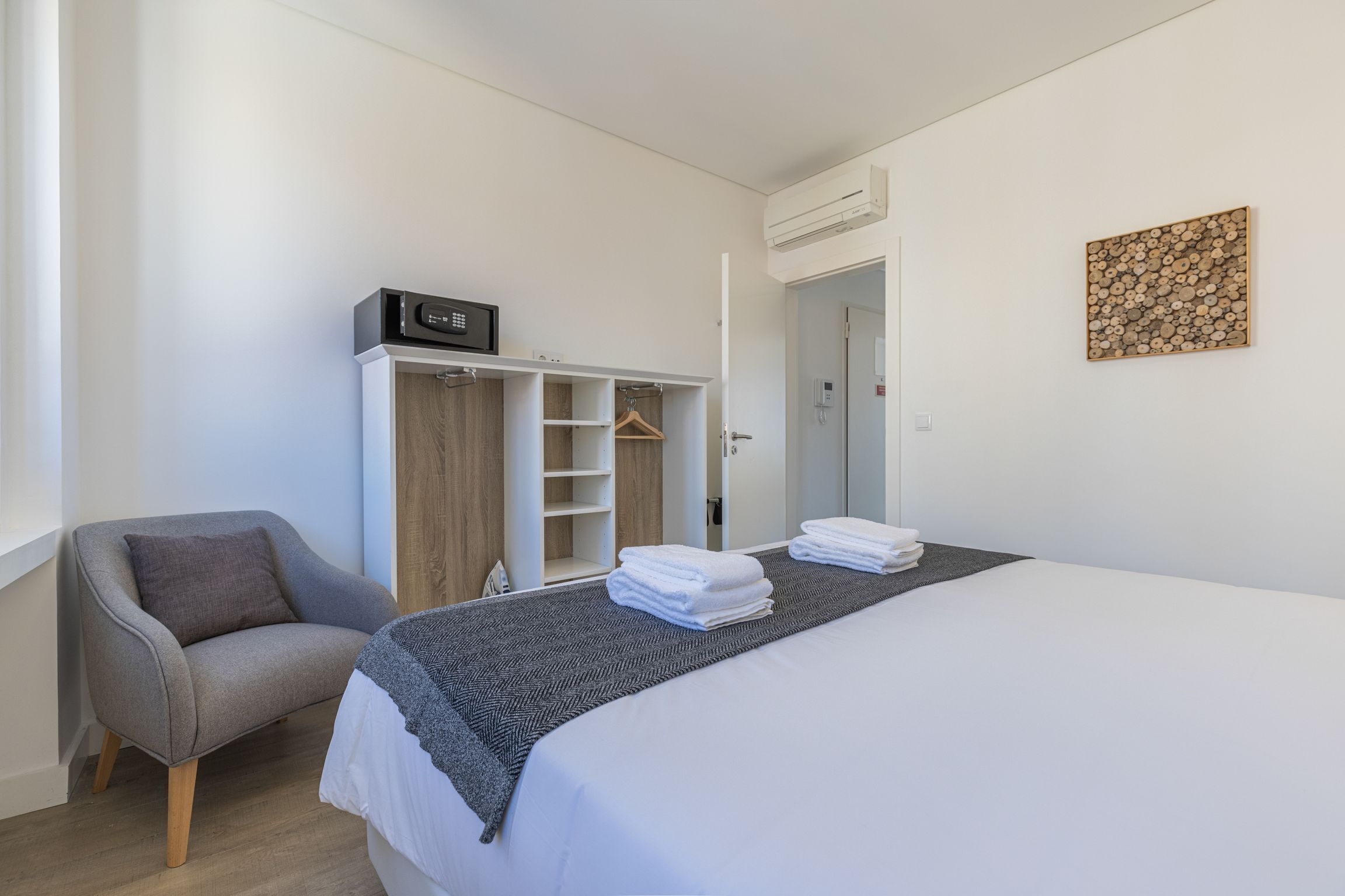 A modern bedroom with a bed, a grey armchair, a wardrobe, and a wall-mounted artwork.