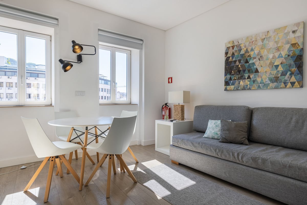 A modern living room with a grey sofa, white chairs, a round table, and a geometric painting on the wall.