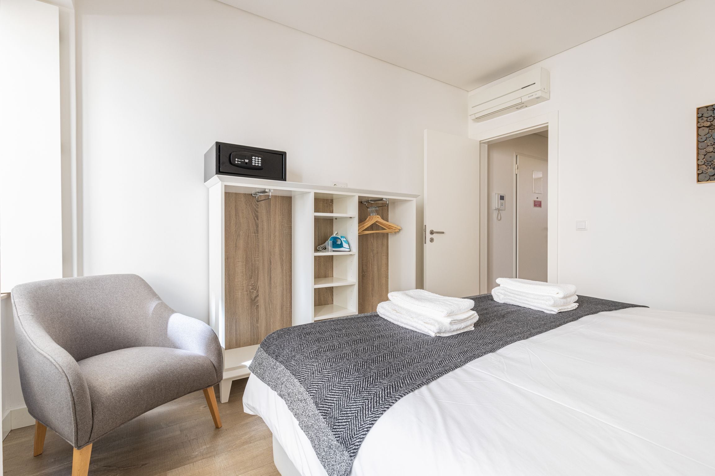 Une chambre d’hôtel avec un lit, un fauteuil gris, une armoire et un manteau accroché à la porte.