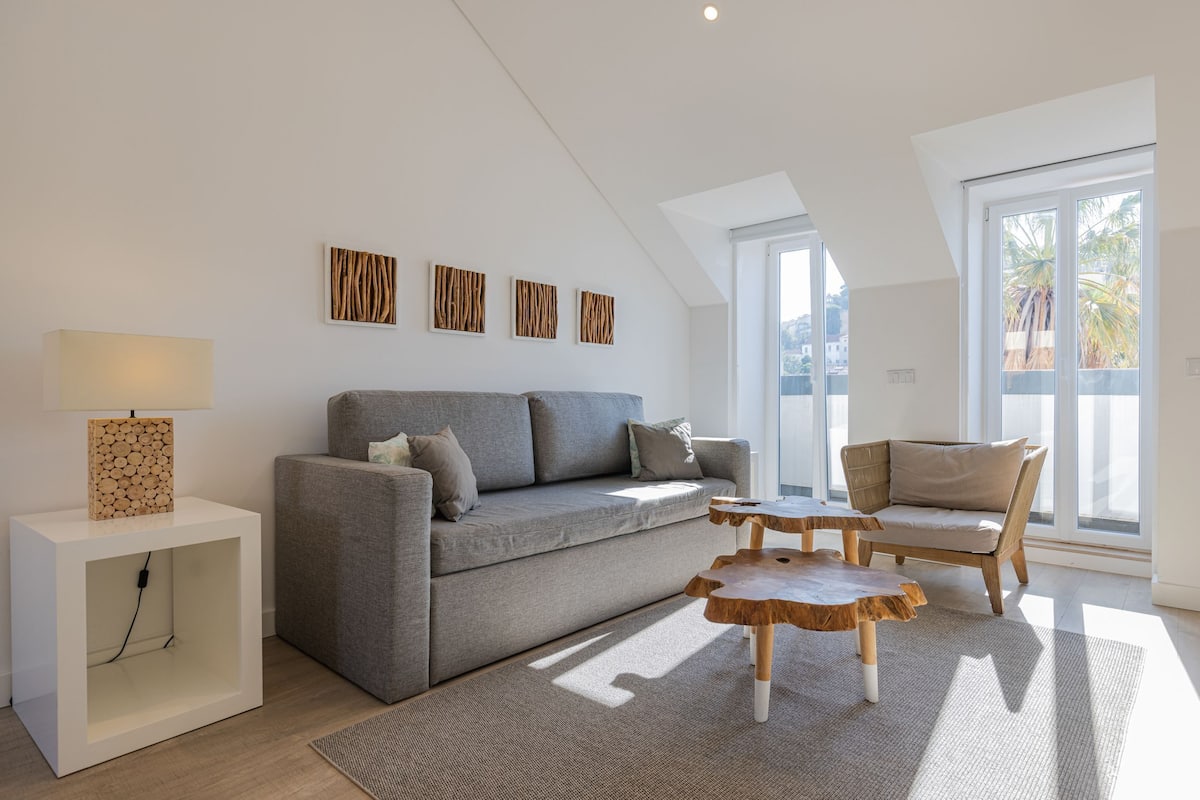 A modern living room with a grey sofa, a wooden coffee table, and a side table with a lamp. There are three wall-mounted artworks above the sofa.