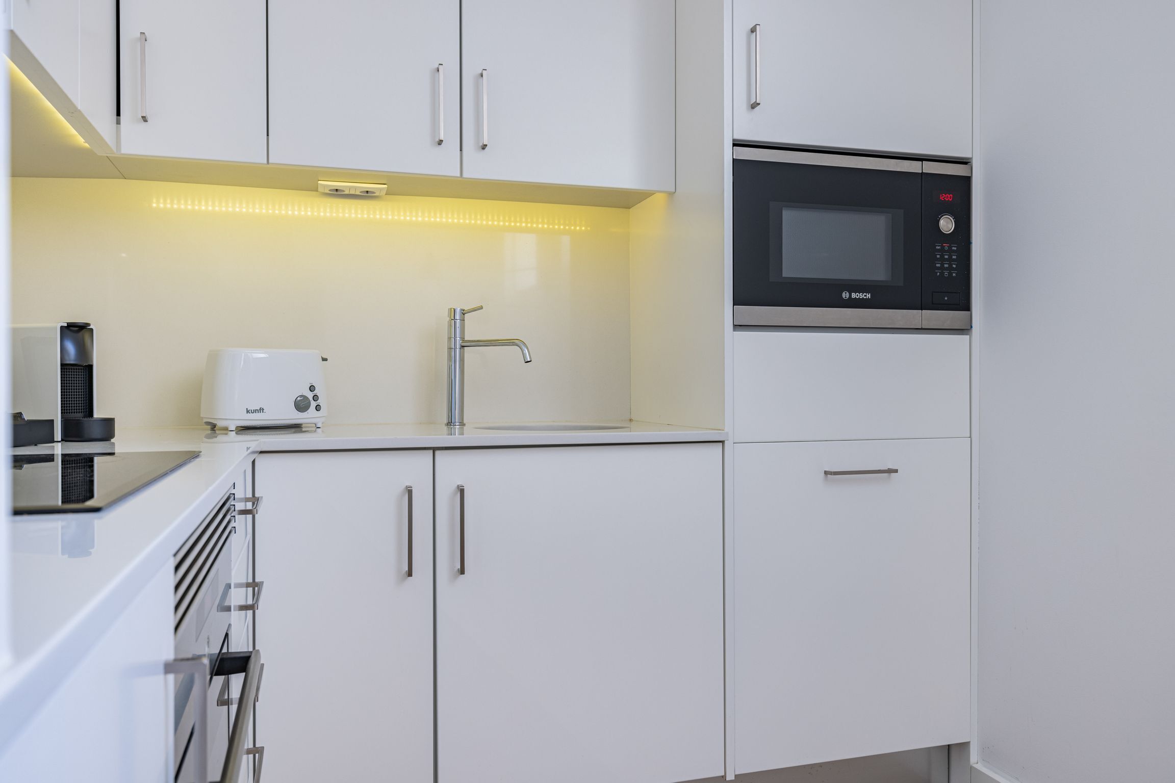 A modern kitchen with white cabinets, a Bosch microwave, a coffee maker, and a built-in oven.