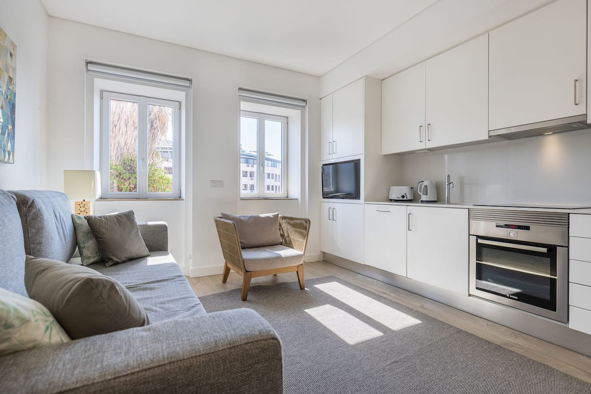 A modern living room with a sofa, armchairs, and a kitchen area with white cabinets and appliances.