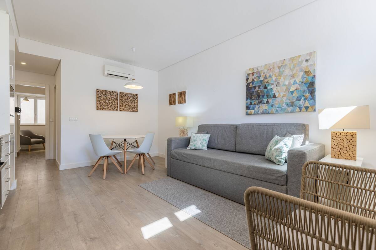A modern living room with a grey sofa, two chairs, a small table, and a wicker chair. There are wall-mounted air conditioning units and a geometric artwork above the sofa.
