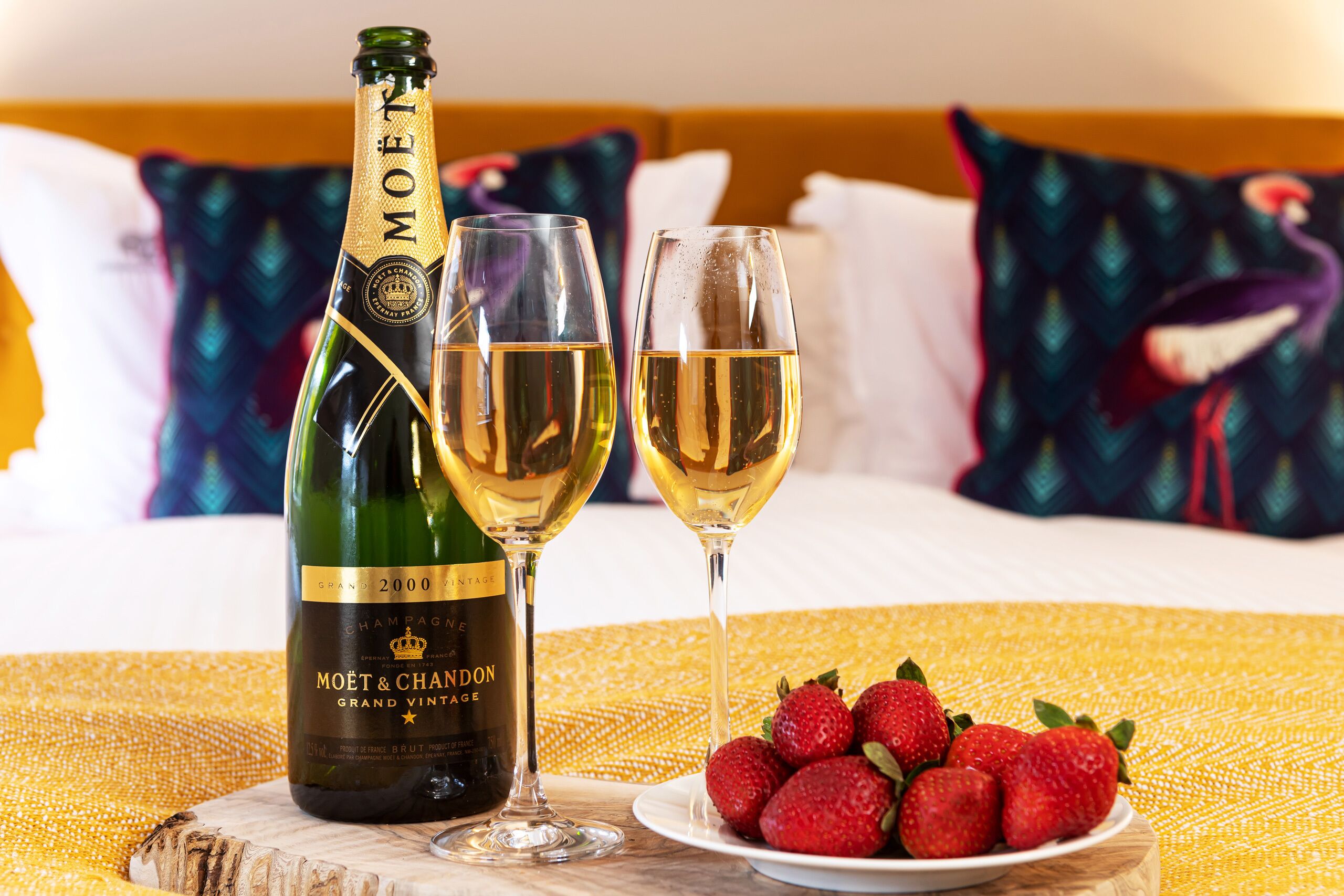Uma garrafa de champanhe Moët & Chandon Grand Vintage, dois copos de champanhe e um prato de morangos sobre uma cama com almofadas ao fundo.