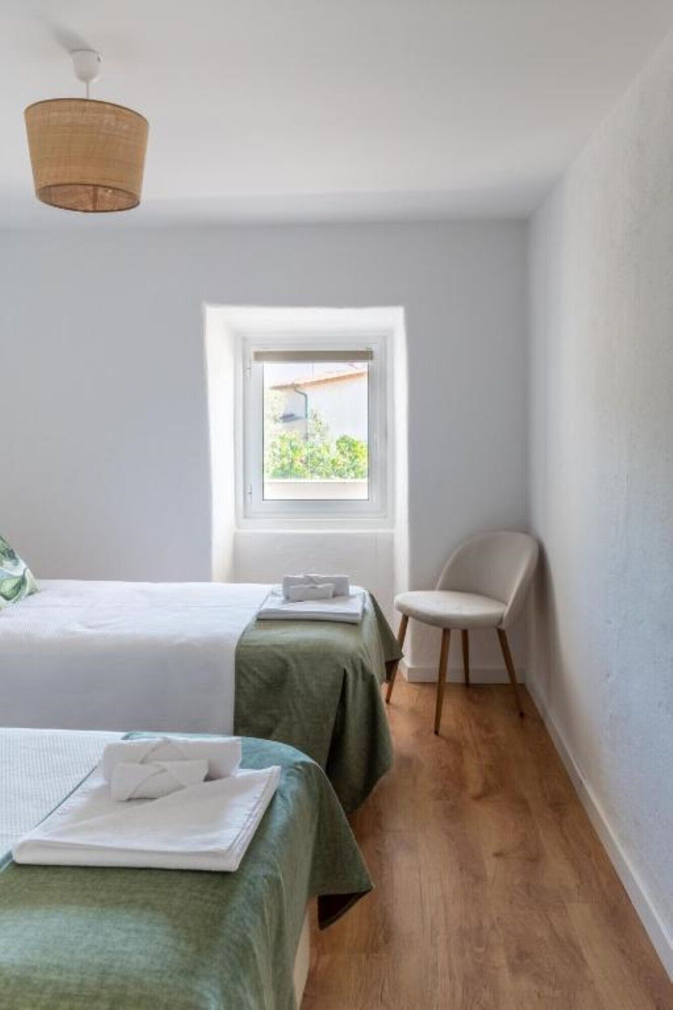 A small, bright room with two beds, a chair, and a window providing a view of greenery outside.