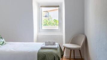 A small, bright room with two beds, a chair, and a window providing a view of greenery outside.