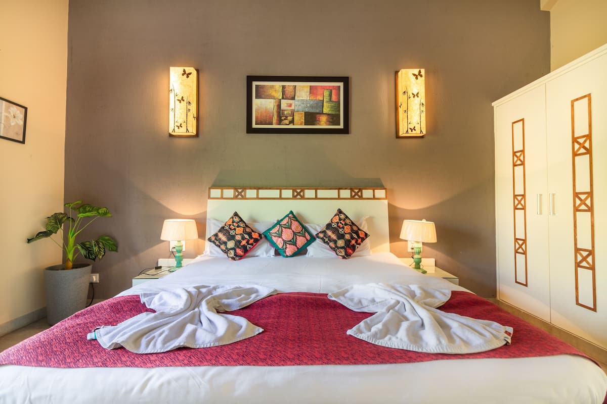 A neatly made bed with a red blanket, two bedside lamps, and a potted plant on the left.
