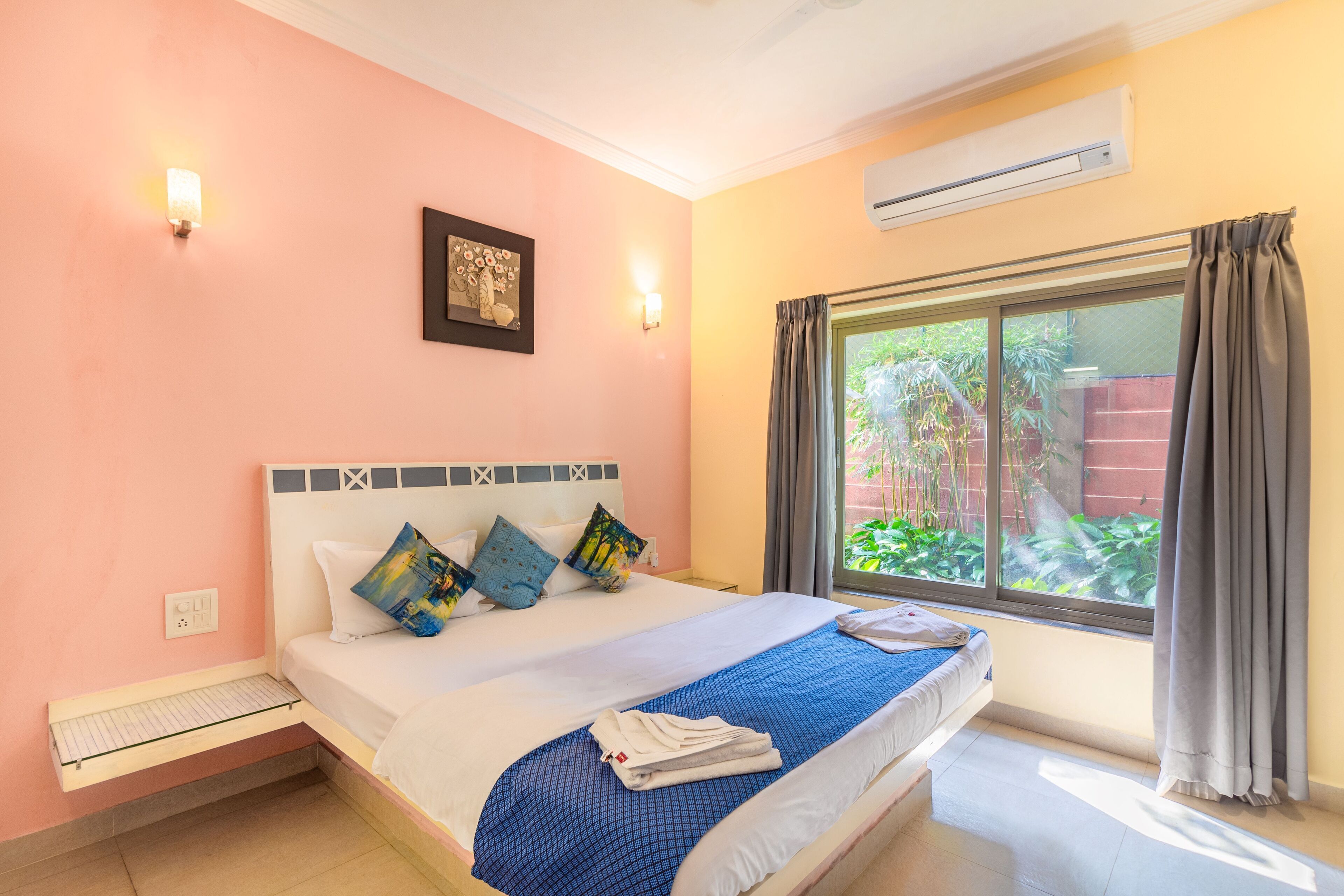 A neatly made bed with a blue bedspread and multiple pillows, a bedside table with a lamp, and a window with curtains.