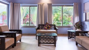 A room with a sofa, two armchairs, a glass-top coffee table, and a small side table. Large windows with curtains provide a view of greenery outside. - Luxury villa Diva Goa (Calangute)