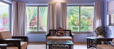 Una habitación con un sofá, dos sillones, una mesa de centro de vidrio y una mesita auxiliar. Ventanas amplias con cortinas ofrecen vistas a zonas verdes al exterior.