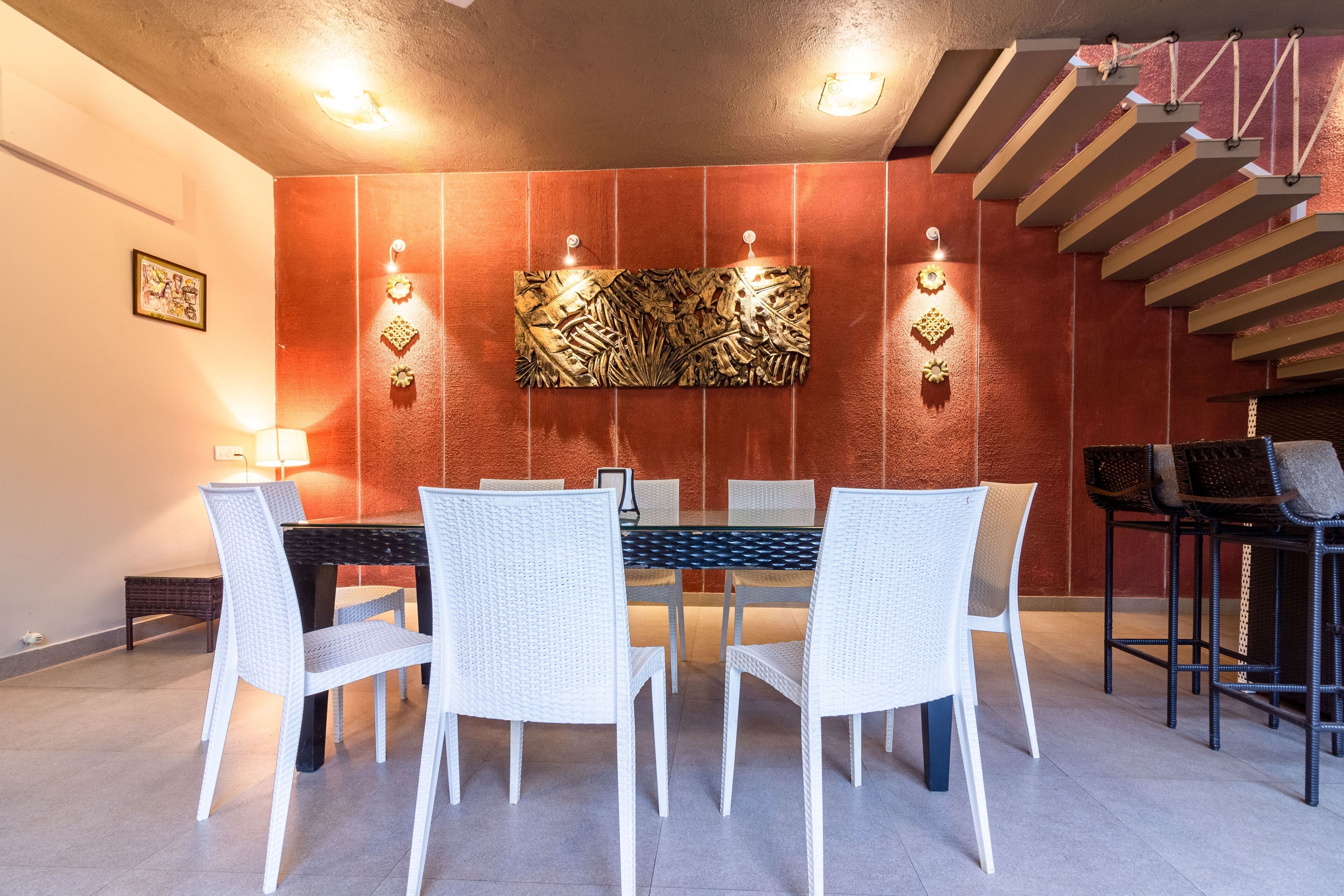 A dining area with a glass-top table surrounded by white chairs, a decorative wall piece, and a staircase in the background.
