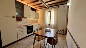 Una cucina compatta con mobili bianchi, zoccolo piastrellato, tavolo da pranzo in legno con sedie e finestra con tende.