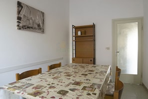 Un coin repas avec une table recouverte d’une nappe à motifs, des chaises en bois, une bibliothèque et une porte.