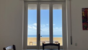 Une pièce avec une table et des chaises blanches, une grande fenêtre donnant sur la plage et l’océan, et un tableau accroché au mur.