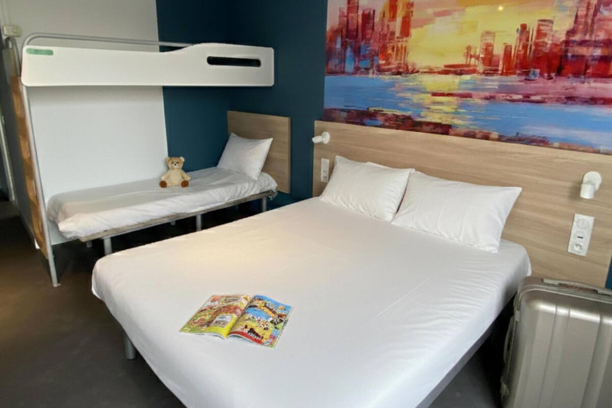 A hotel room with a large bed, a smaller bed, a wall-mounted TV, a painting, and a suitcase.