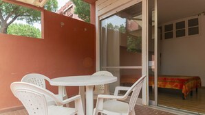Een terras met een witte tafel en vier stoelen, een schuifdeur die leidt naar een kamer met een bed, en een rode muur.