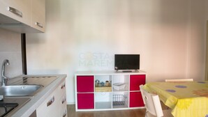 Een keuken met een spoelbak, fornuis en kastjes. Er staat een televisie op een witte kast en een tafel met een gele tafelkleed.