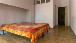 Een kamer met een bed, houten vloer, een witte muur met een raam, en een deur naar een andere kamer.