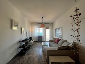 Ein modernes Wohnzimmer mit einer Couch, einem Couchtisch und einem Fernseher. Ein Balkon bietet Blick aufs Meer.