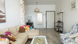 Ett vardagsrum med en soffa, ett soffbord med en glasskiva, en fåtölj med ett blommönster, en hängande växt och en lampa.