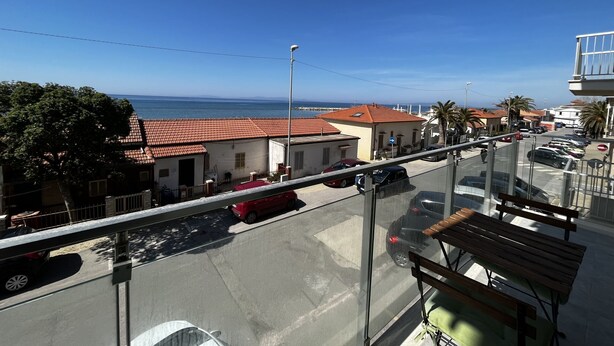 A balcony with a view of the sea, a parking area, and a building with a red-tiled roof. - SPIAGGIA DI LEVANTE - ELBA (Follonica)