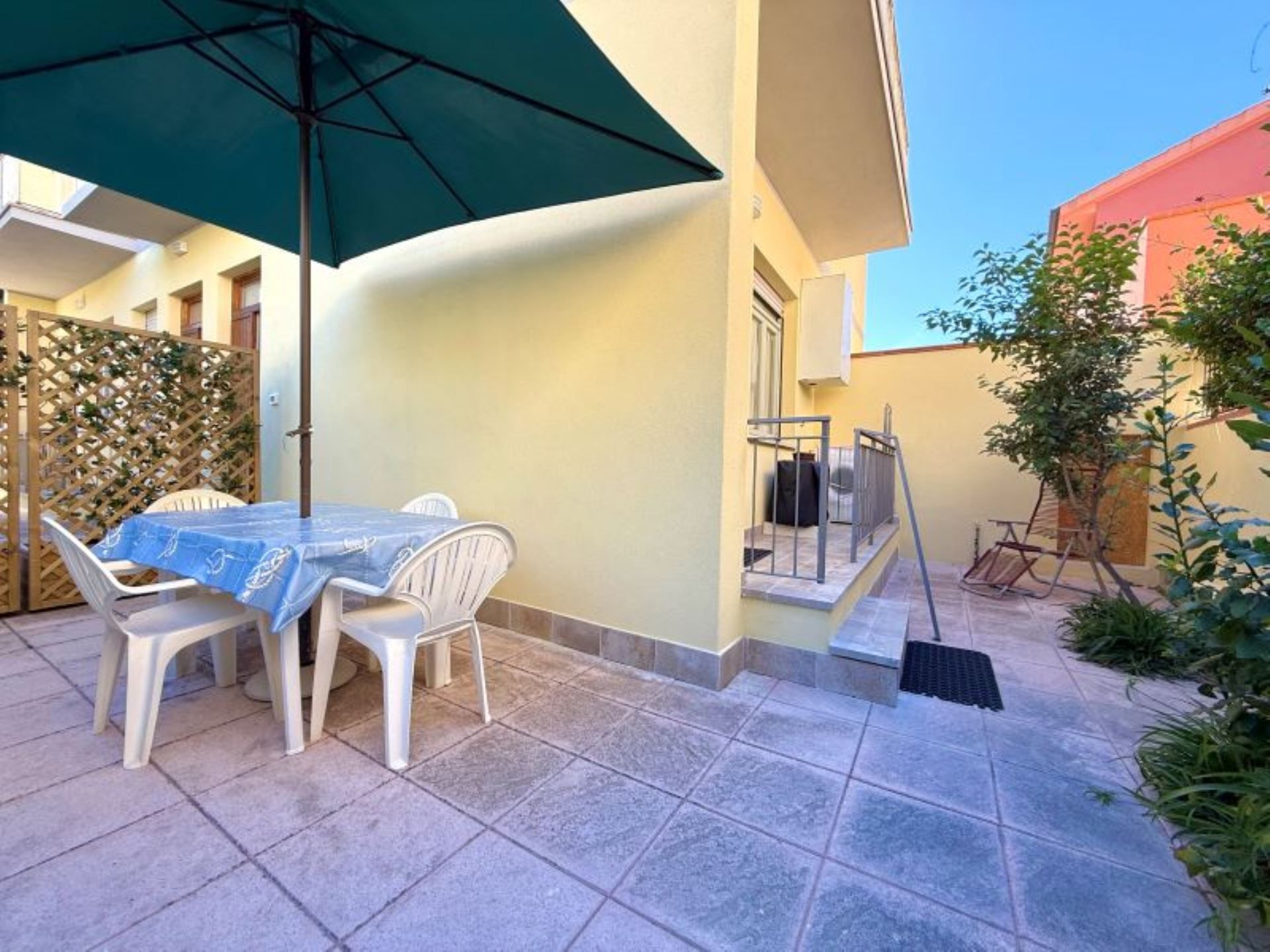 Eine Terrassengruppe im Freien mit Tisch, Stühlen und einem grünen Sonnenschirm, sowie eine Treppe zu einem Balkon.