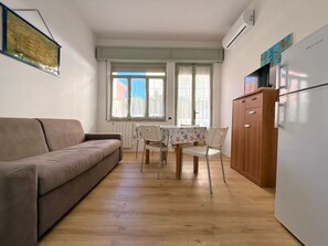 A compact living space with a sofa, dining table, chairs, and a refrigerator. - GIANNUTRI - NODO SAVOIA (Follonica)