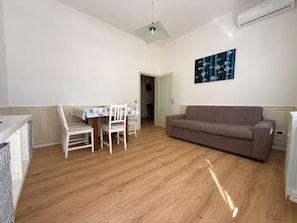 Ein Zimmer mit Holzboden, einem braunen Sofa, einem Esstisch mit Stühlen und einer Wand-Klimaanlage.