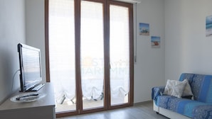 Una sala de estar moderna con un televisor de pantalla plana sobre una mesa de madera, un sofá azul con estampado y un amplio ventanal corredero con vista a la playa.