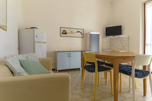 Ein kompakter Wohnraum mit einer Couch, einem kleinen Esstisch mit Stühlen, einer Küchenzeile mit Kühlschrank, einem Fernseher und einem gerahmten Bild an der Wand.