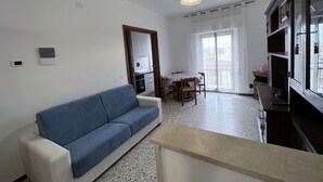 A living room with a blue sofa, a dining table with chairs, a kitchen area, and a large window with curtains. - Salvia apartment in Follonica (Follonica)