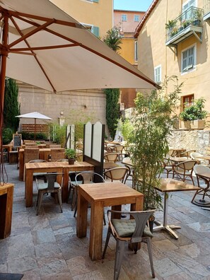 An outdoor café with wooden tables and chairs, covered by large umbrellas, set against a backdrop of buildings and greenery. - LE MAY AP4338 By Riviera Holiday Homes (Villefranche-sur-Mer)