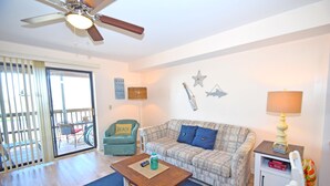A living room with a ceiling fan, a sofa, a coffee table, a lamp, and a small cabinet. - Raffles 206-Bayside 127th St, Free WiFi, Pool, W/D, AC (Ocean City)
