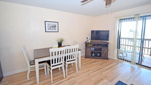 A dining area with a wooden table and white chairs, a ceiling fan, a television on a wooden stand, and a window with a view of a deck. - Raffles 206-Bayside 127th St, Free WiFi, Pool, W/D, AC (Ocean City)