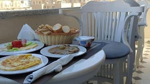 Un balcón con una mesa dispuesta para el desayuno, que incluye platos de comida, una cesta de pan y un recipiente con yogur.