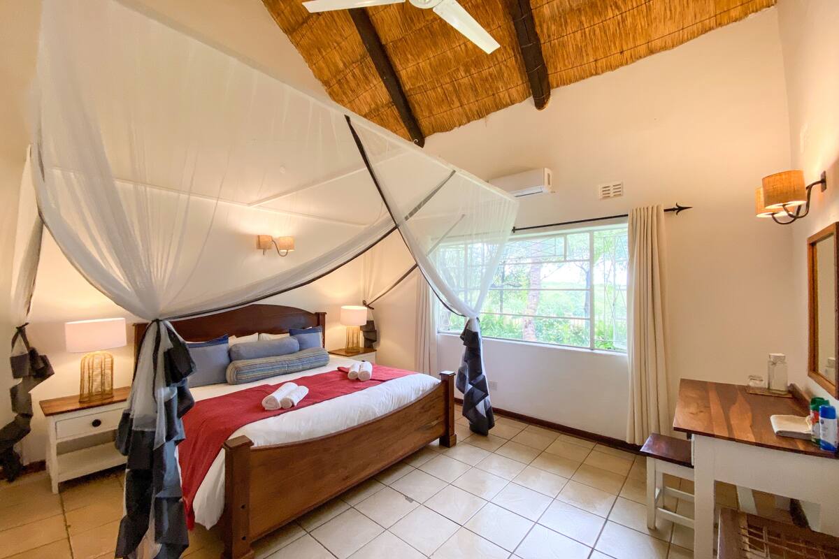 A bedroom with a wooden bed, a ceiling fan, a mosquito net, a window with curtains, a bedside table with a lamp, and a small table with a chair.