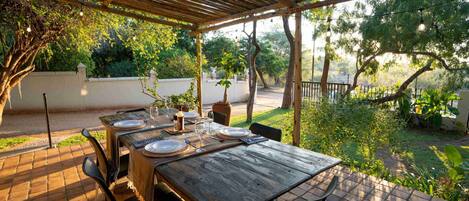 Un patio exterior con una mesa de madera dispuesta para cuatro personas, rodeado de sillas, bajo una pérgola con luces colgantes.