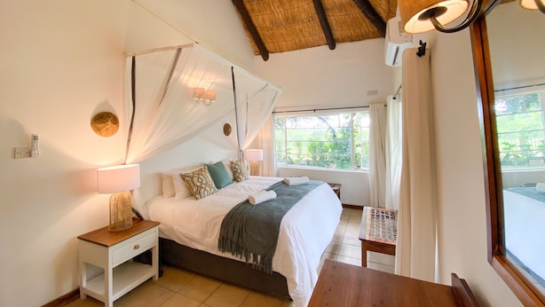 A bedroom with a bed, bedside table, lamp, and a view of greenery outside. - Drift Inn (Victoria Falls)