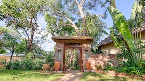 A traditional gate with a thatched roof, surrounded by lush greenery and a paved pathway leading to a building with a thatched roof. - Drift Inn (Victoria Falls)