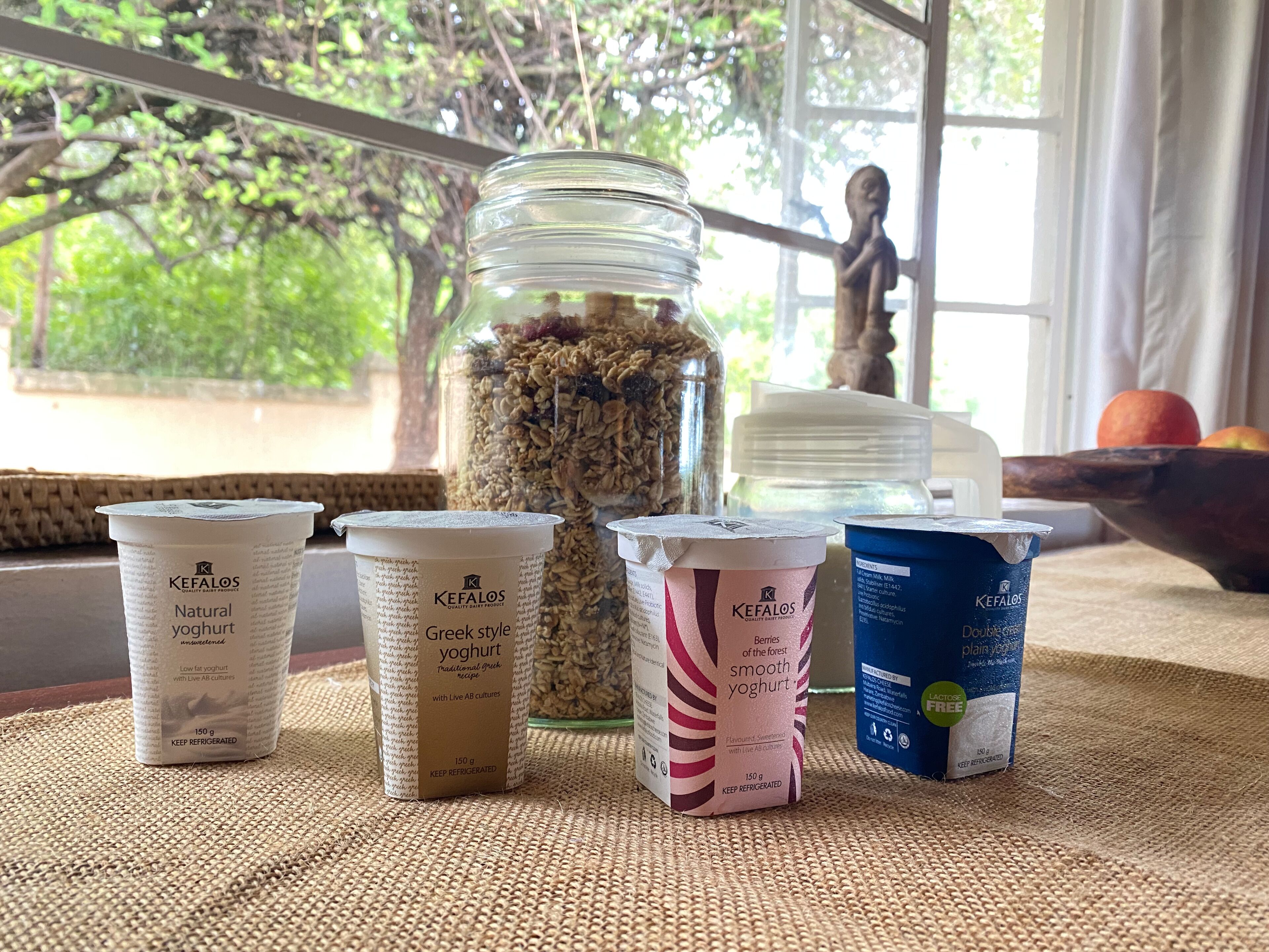 Kefalos natural yogurt, Greek style yogurt, and plain yogurt on a table with a view of greenery outside.