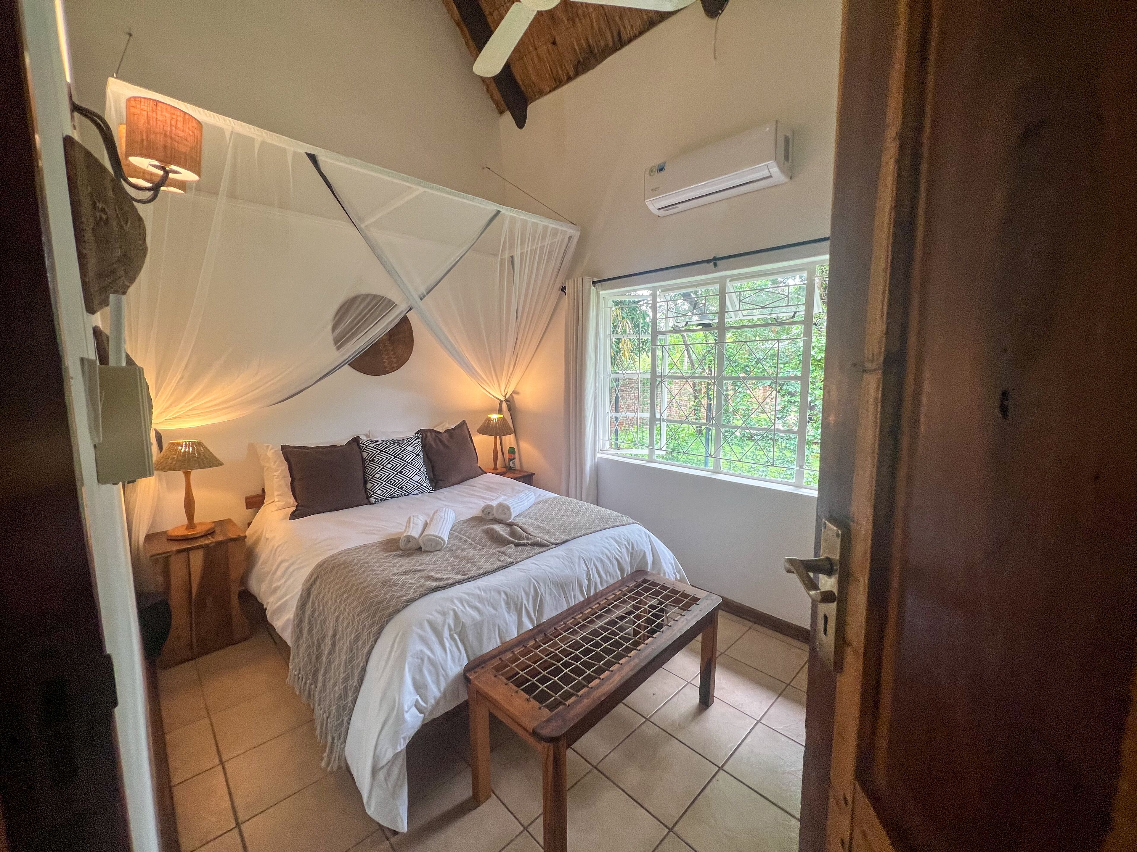 A bedroom with a bed, bedside lamps, a mosquito net, a window with a view, and a wooden bench.