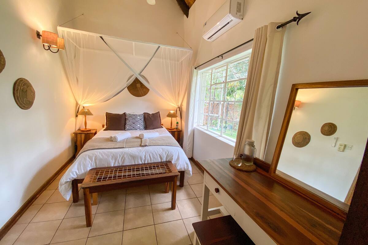 A bedroom with a bed, bedside lamps, a bench, a window, and a wall-mounted air conditioner.