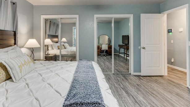 A bedroom with a bed, bedside lamps, a ceiling fan, a mirror, and a door leading to another room.