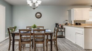 A dining area with a wooden table and chairs, a chandelier, and a kitchen with white cabinets and a countertop.