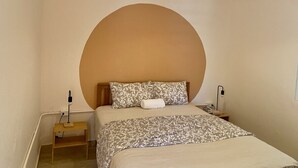 Un dormitorio con un cabecero grande y redondo, una cama con ropa de cama estampada, una mesita de noche con una lámpara y un equipo de aire acondicionado en la pared.