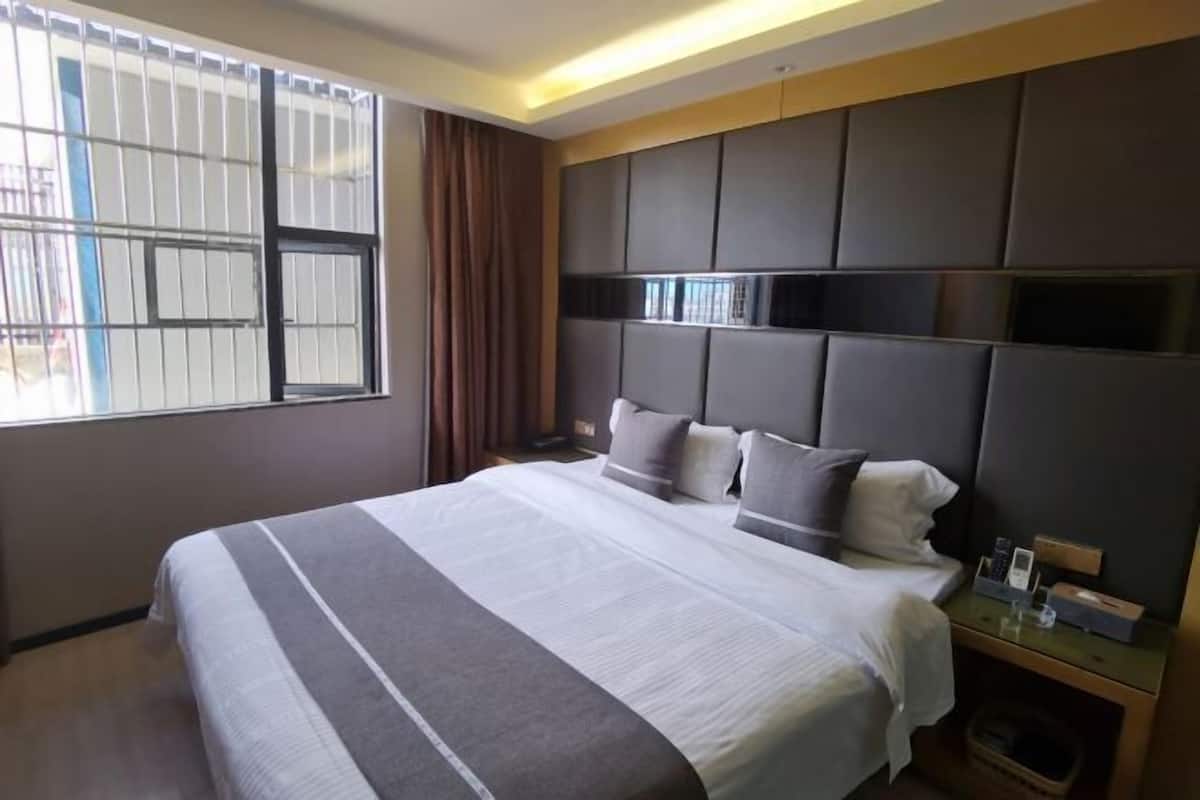 A hotel room with a large bed, a headboard with built-in shelves, a bedside table with a phone and a lamp, and a window with bars.