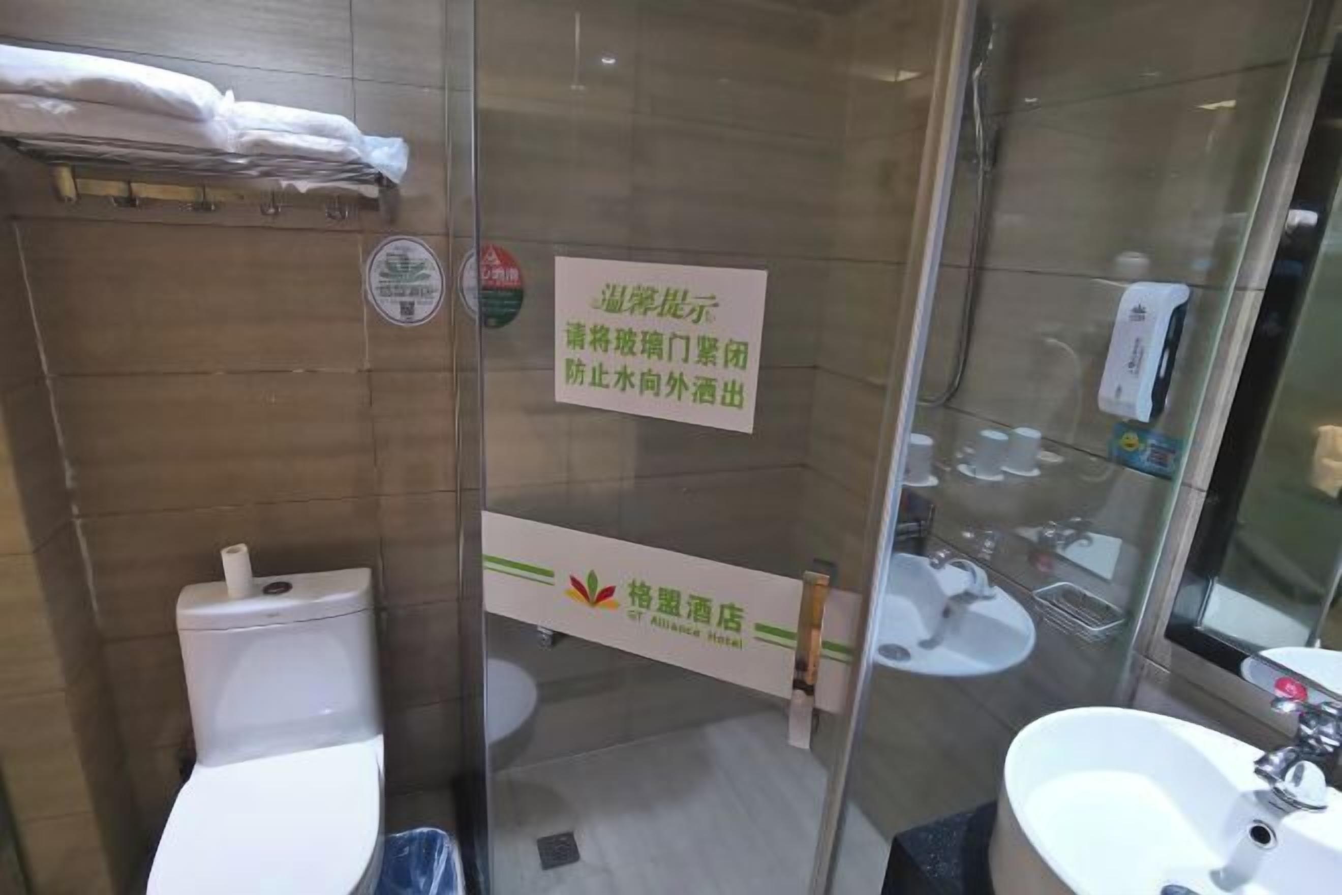 Un baño de hotel con separador de vidrio, inodoro, lavabo y zona de ducha. Hay un letrero en el separador de vidrio con texto en un idioma extranjero.