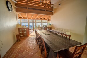 Un vaste espace dédié à la salle à manger, avec une longue table en bois et des chaises, une horloge au mur et une vue sur la mer grâce à de grandes fenêtres.